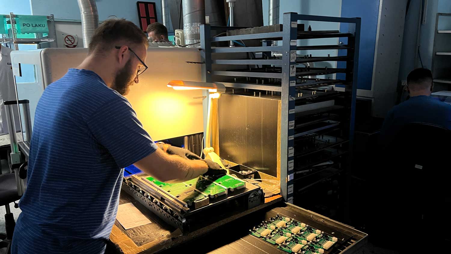 A technician assembling circuit boards at a production workstation, with populated boards racked beside the fixture.