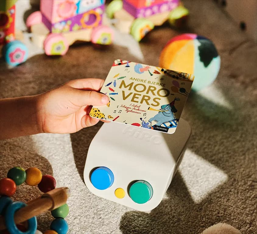 A child's hand holding a story card above the HiRO speaker while playing among toys on the floor.