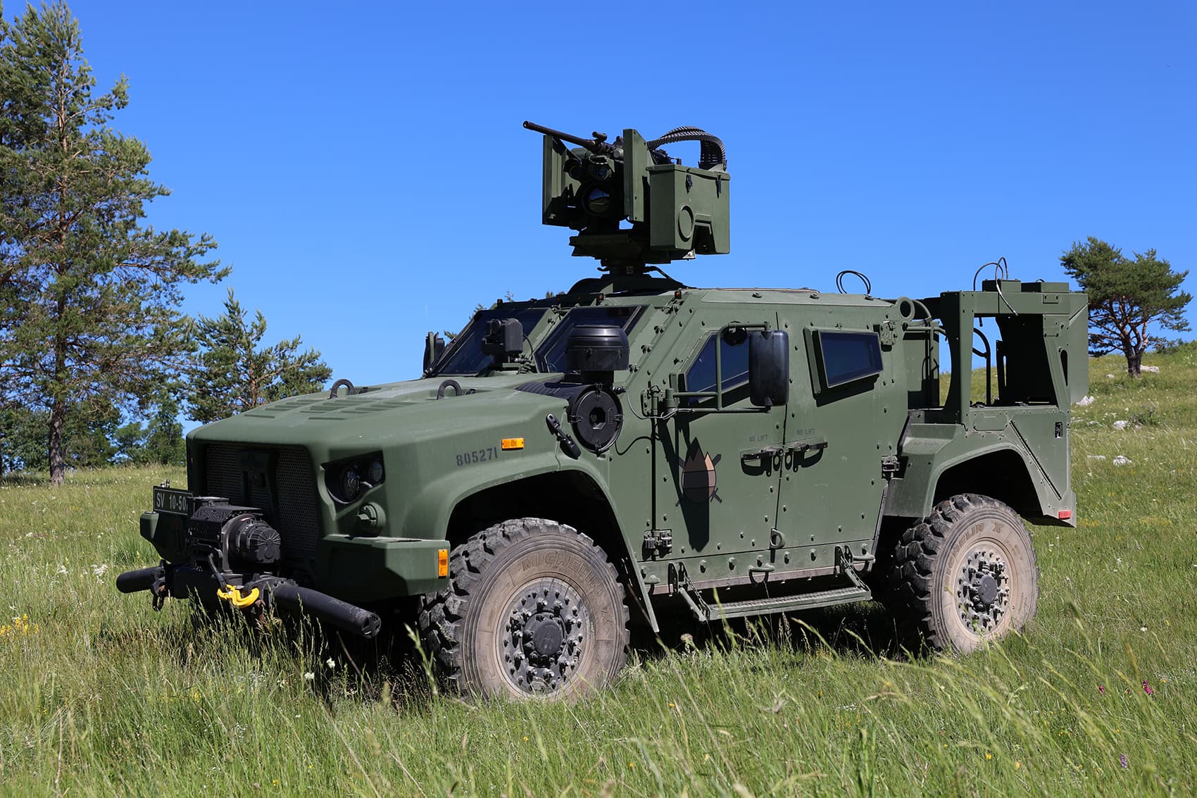 An armored military vehicle fitted with a remote weapon station, parked in a grass field.