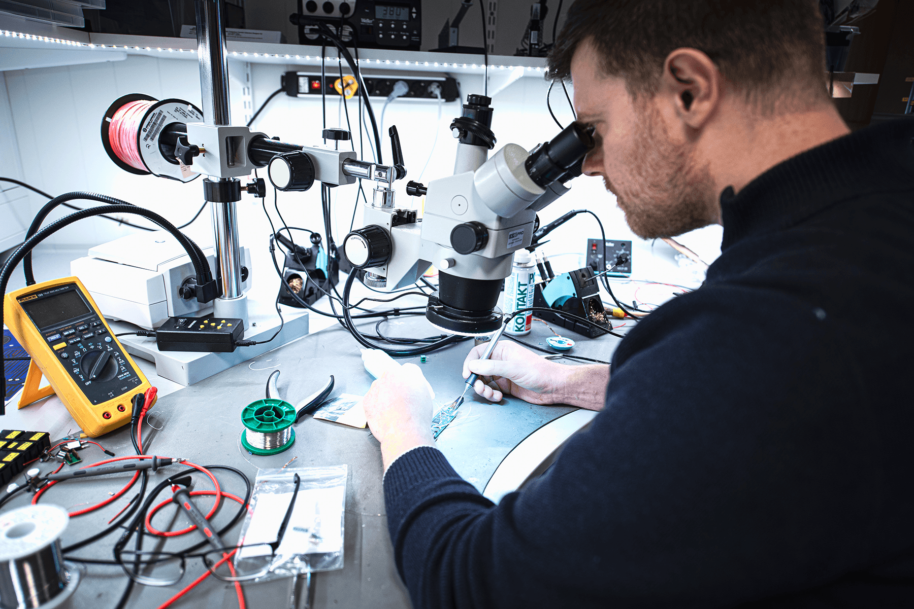 An engineer solders a small circuit board while viewing it through a microscope, with a multimeter and wire spools on the workbench.